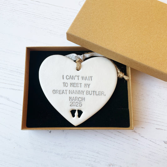 Pearlised white clay hanging heart with baby feet cut out of the bottom and grey personalisation, the heart is personalised with I CAN’T WAIT TO MEET MY GREAT NANNY BUTLER. MARCH 2025