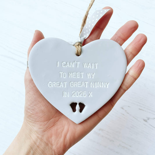 Grey clay hanging heart with baby feet cut out of the bottom and white personalisation, the heart is personalised with I CAN’T WAIT TO MEET MY GREAT GREAT NINNY IN 2025 X
