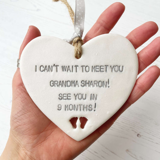 Pearlised white clay hanging heart pregnancy reveal with baby feet cut out of the bottom and grey personalisation, the heart is personalised with I CAN’T WAIT TO MEET YOU GRANDMA! SEE YOU IN 9 MONTHS!