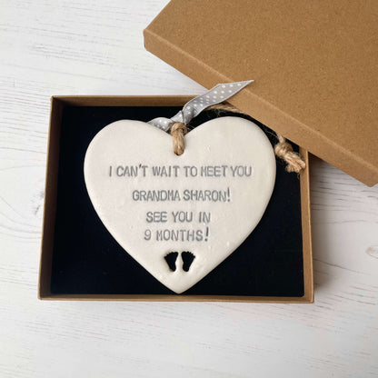 Pearlised white clay hanging heart pregnancy reveal with baby feet cut out of the bottom and grey personalisation, the heart is personalised with I CAN’T WAIT TO MEET YOU GRANDMA! SEE YOU IN 9 MONTHS!