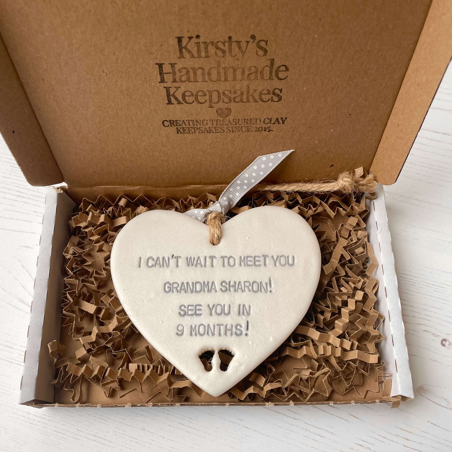 Pearlised white clay hanging heart pregnancy reveal with baby feet cut out of the bottom and grey personalisation, the heart is personalised with I CAN’T WAIT TO MEET YOU GRANDMA! SEE YOU IN 9 MONTHS!