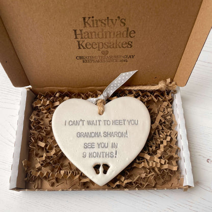 Pearlised white clay hanging heart pregnancy reveal with baby feet cut out of the bottom and grey personalisation, the heart is personalised with I CAN’T WAIT TO MEET YOU GRANDMA! SEE YOU IN 9 MONTHS!