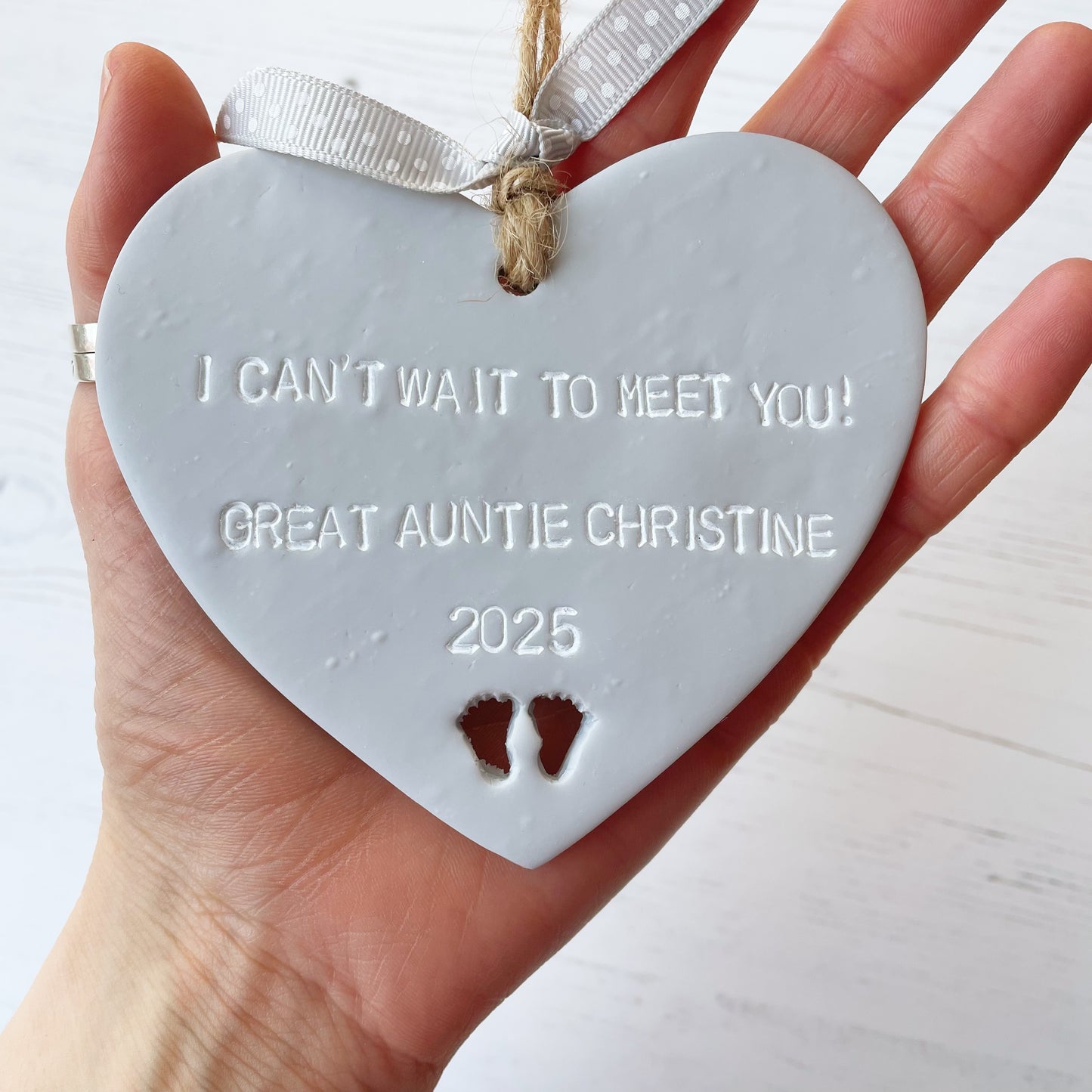 Grey clay hanging heart with baby feet cut out of the bottom and white personalisation, the heart is personalised with I CAN’T WAIT TO MEET YOU! GREAT AUNTIE CHRISTINE 2025