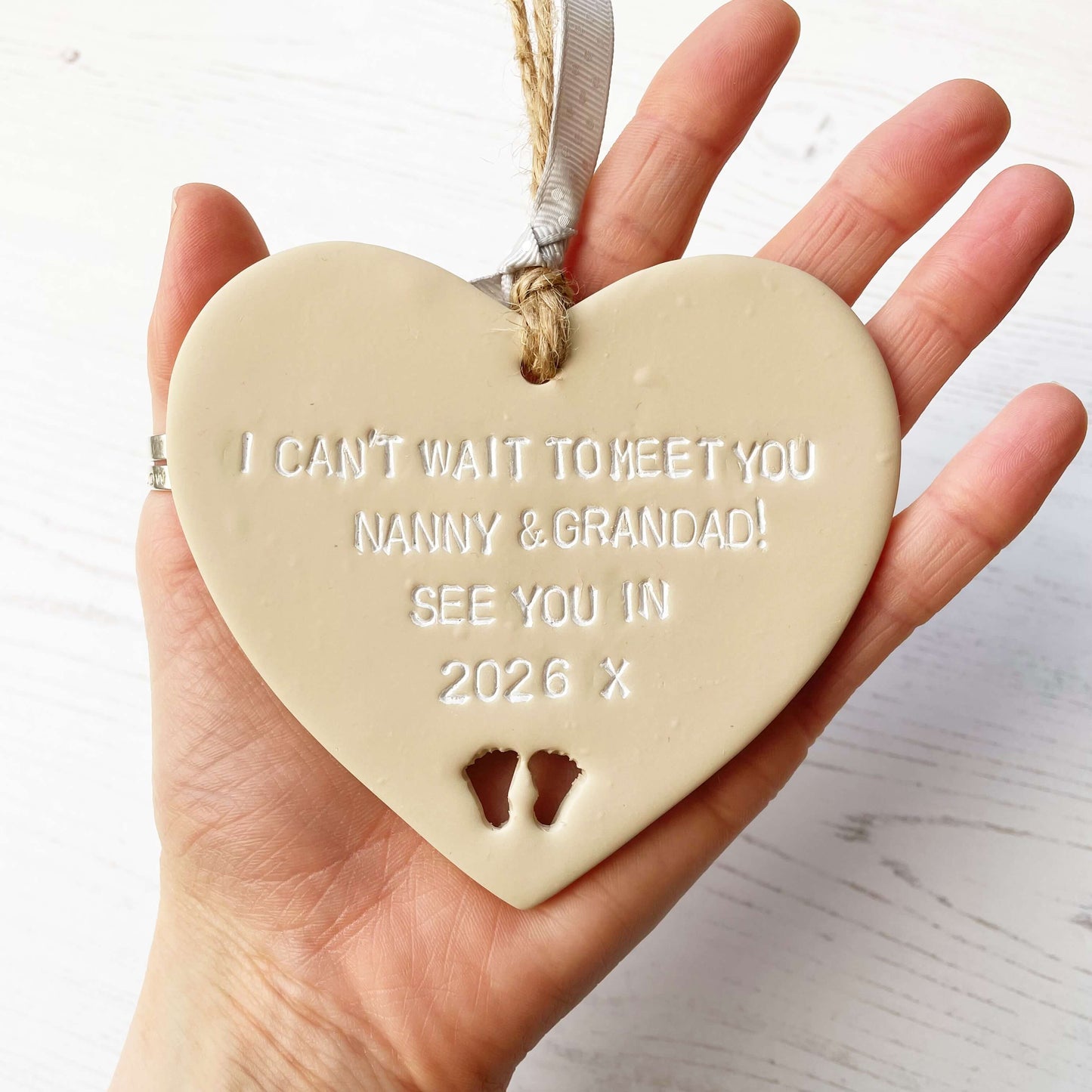 Beige clay hanging heart with baby feet cut out of the bottom and white personalisation, the heart is personalised with I CAN’T WAIT TO MEET MY NANNY & GRANDAD SEE YOU IN 2026