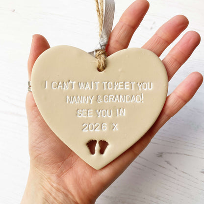 Beige clay hanging heart with baby feet cut out of the bottom and white personalisation, the heart is personalised with I CAN’T WAIT TO MEET MY NANNY & GRANDAD SEE YOU IN 2026