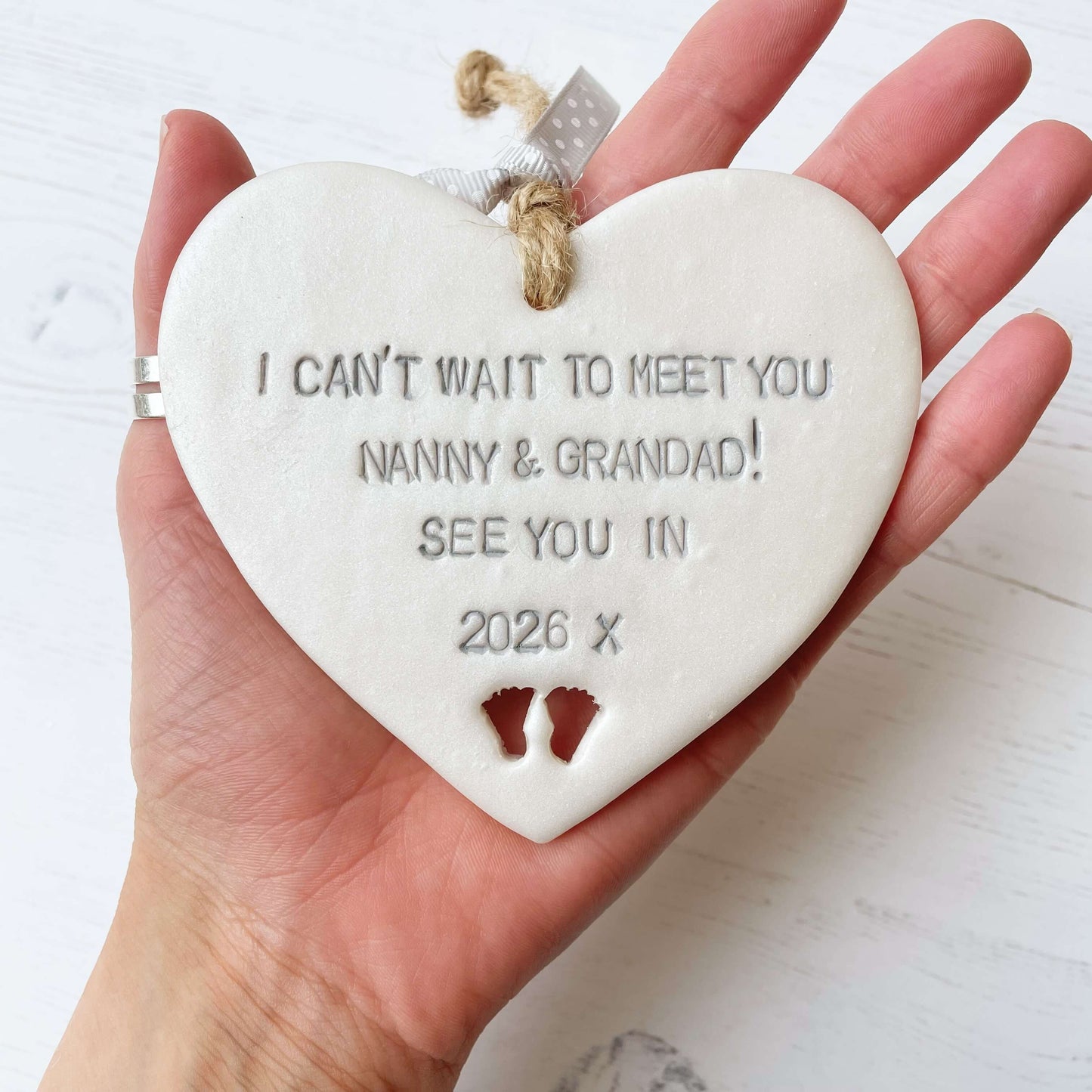 Pearlised white clay hanging heart with baby feet cut out of the bottom and grey personalisation, the heart is personalised with I CAN’T WAIT TO MEET YOU NANNY & GRANDAD! SEE YOU IN 2026 X