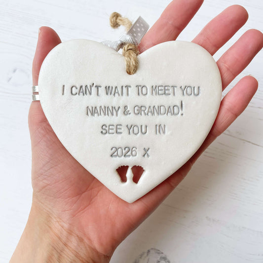 Pearlised white clay hanging heart with baby feet cut out of the bottom and grey personalisation, the heart is personalised with I CAN’T WAIT TO MEET YOU NANNY & GRANDAD! SEE YOU IN 2026 X