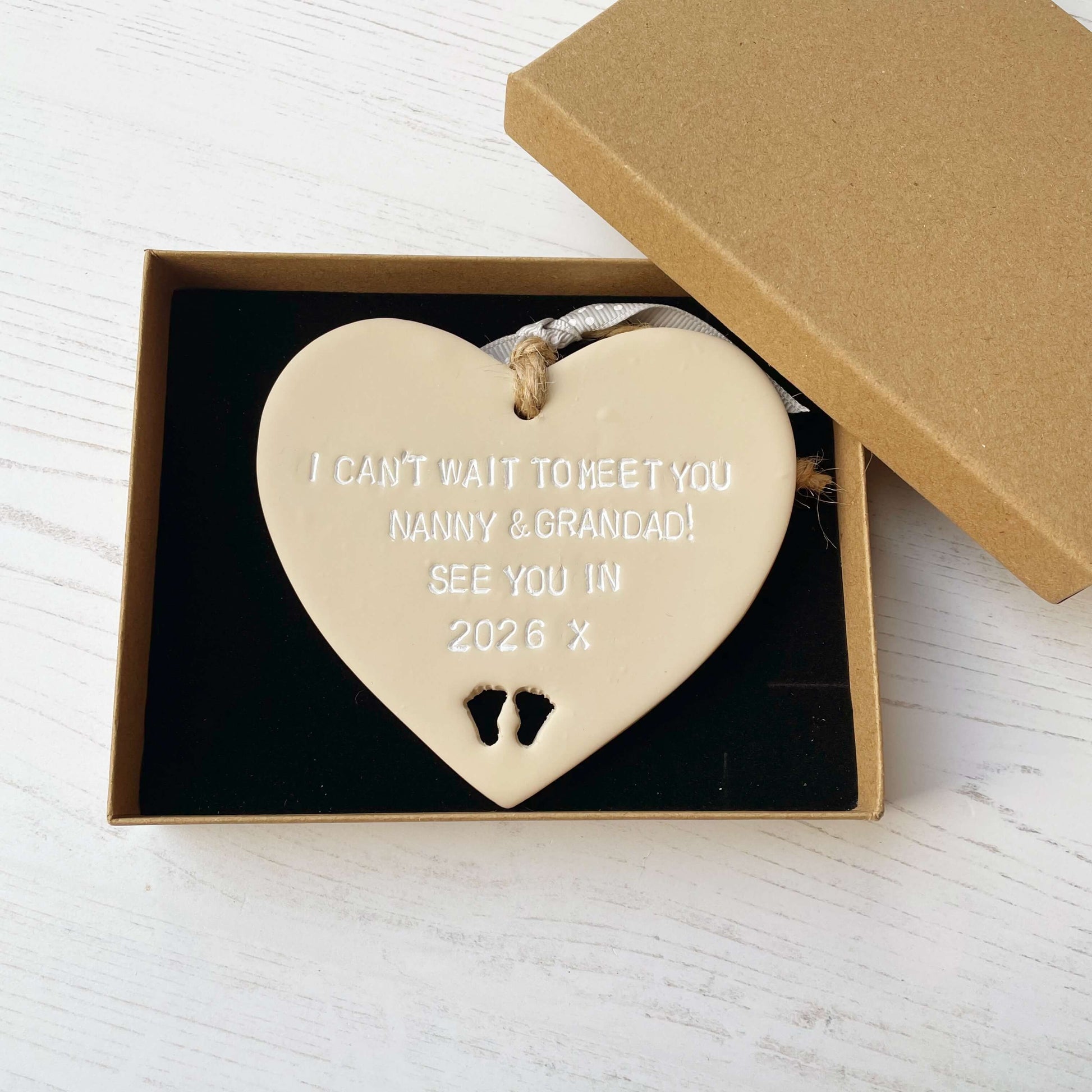 Beige clay hanging heart with baby feet cut out of the bottom and white personalisation, the heart is personalised with I CAN’T WAIT TO MEET MY NANNY & GRANDAD SEE YOU IN 2026