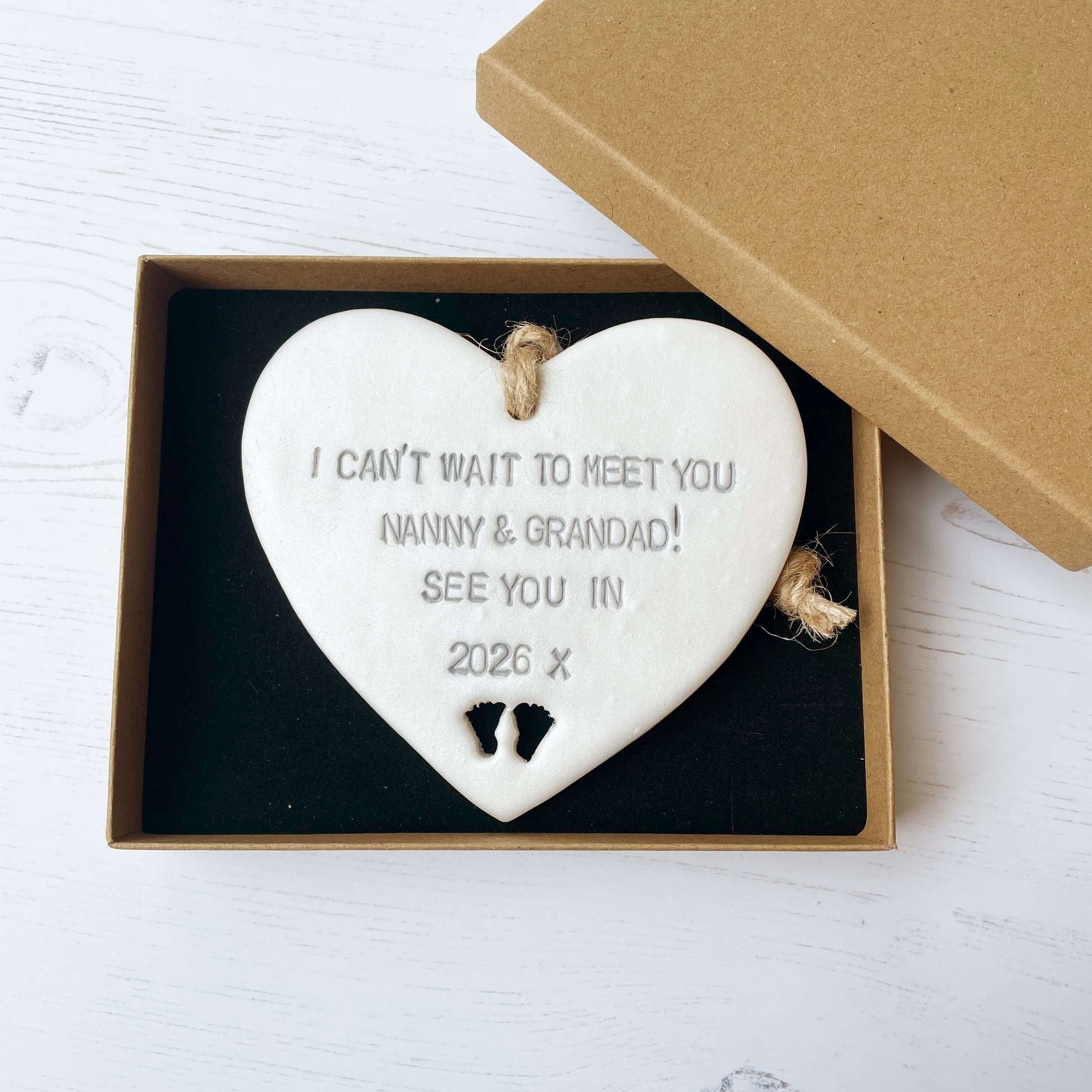 Pearlised white clay hanging heart with baby feet cut out of the bottom and grey personalisation, the heart is personalised with I CAN’T WAIT TO MEET YOU NANNY & GRANDAD! SEE YOU IN 2026 X