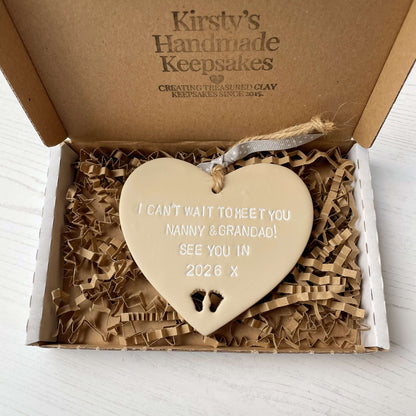 Beige clay hanging heart with baby feet cut out of the bottom and white personalisation, the heart is personalised with I CAN’T WAIT TO MEET MY NANNY & GRANDAD SEE YOU IN 2026
