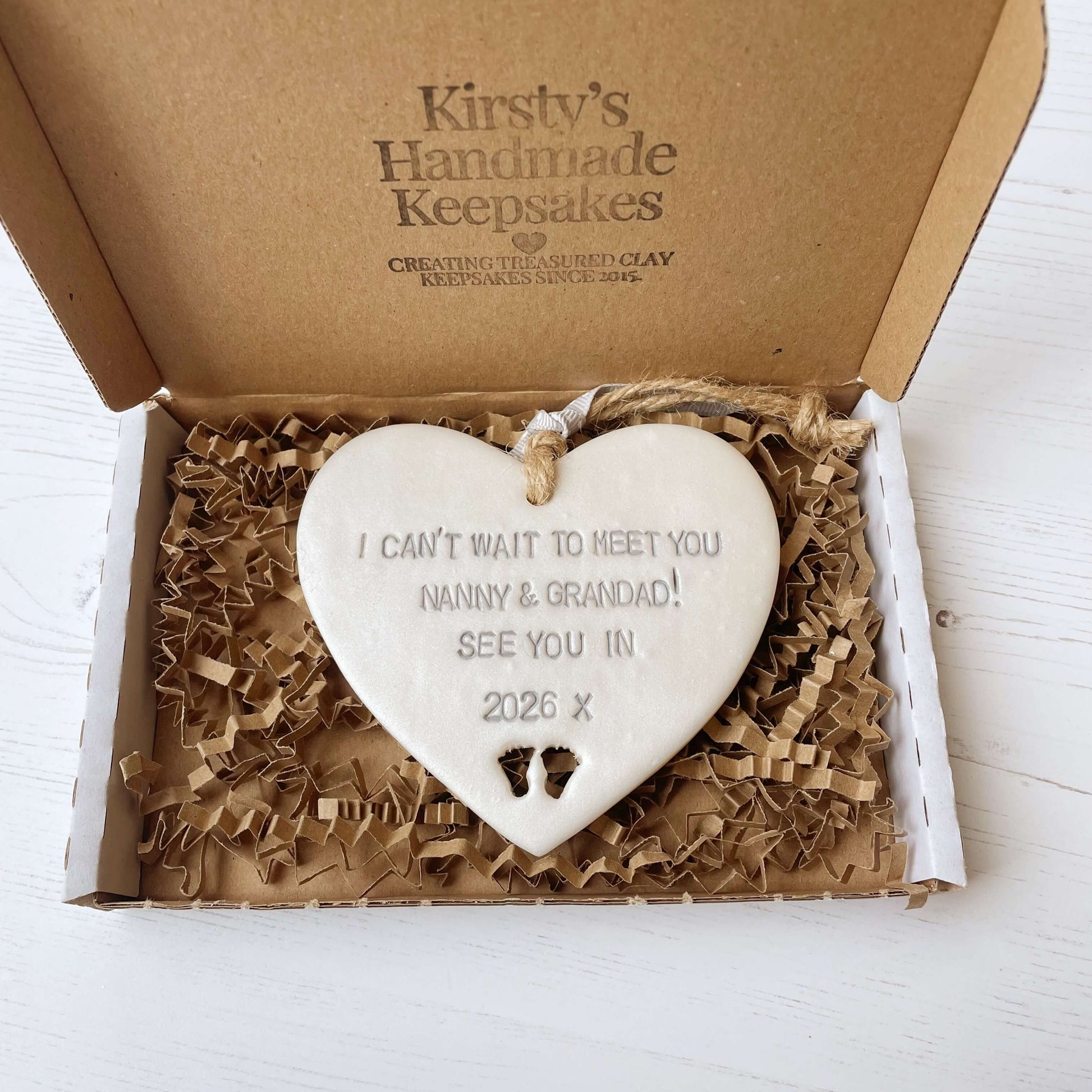 Pearlised white clay hanging heart with baby feet cut out of the bottom and grey personalisation, the heart is personalised with I CAN’T WAIT TO MEET YOU NANNY & GRANDAD! SEE YOU IN 2026 X
