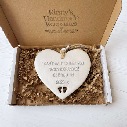 Pearlised white clay hanging heart with baby feet cut out of the bottom and grey personalisation, the heart is personalised with I CAN’T WAIT TO MEET YOU NANNY & GRANDAD! SEE YOU IN 2026 X