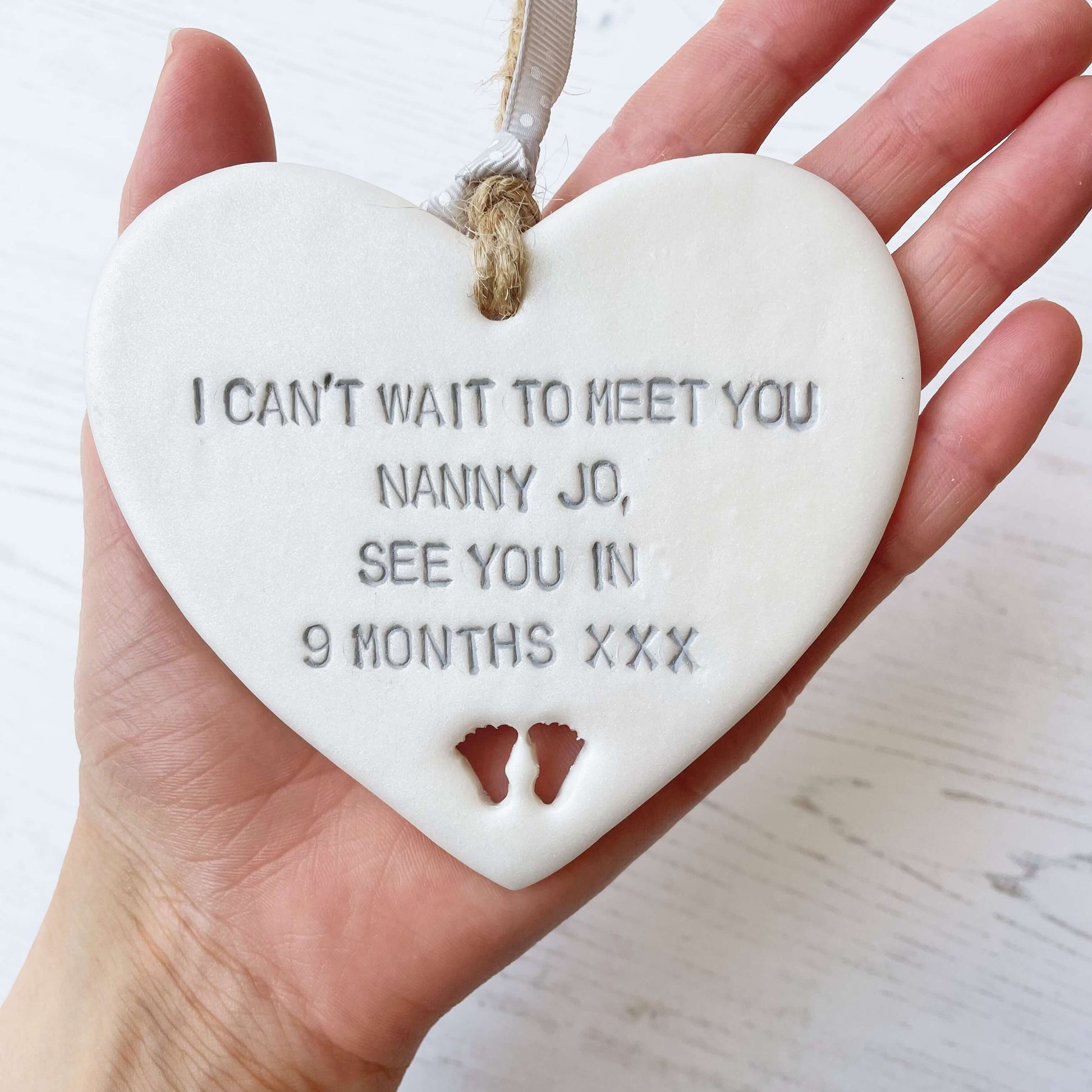 Pearlised white clay hanging heart with baby feet cut out of the bottom and grey personalisation, the heart is personalised with I CAN’T WAIT TO MEET YOU NANNY JO, SEE YOU IN 9 MONTHS XXX