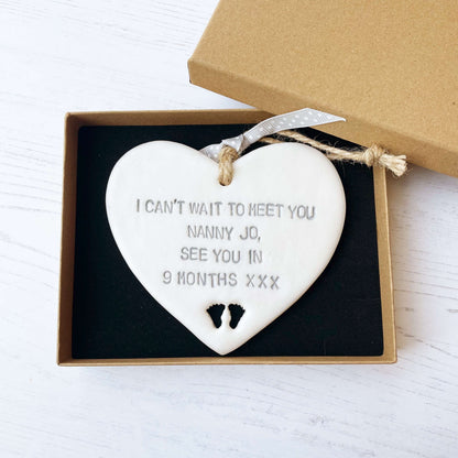 Pearlised white clay hanging heart with baby feet cut out of the bottom and grey personalisation, the heart is personalised with I CAN’T WAIT TO MEET YOU NANNY JO, SEE YOU IN 9 MONTHS XXX