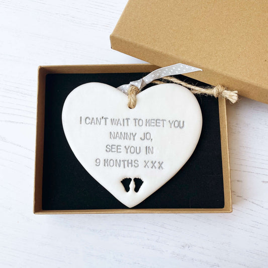 Pearlised white clay hanging heart with baby feet cut out of the bottom and grey personalisation, the heart is personalised with I CAN’T WAIT TO MEET YOU NANNY JO, SEE YOU IN 9 MONTHS XXX