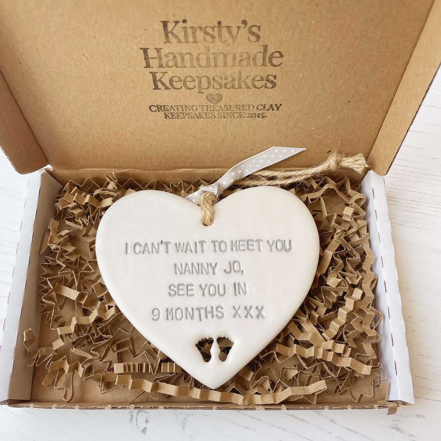 Pearlised white clay hanging heart with baby feet cut out of the bottom and grey personalisation, the heart is personalised with I CAN’T WAIT TO MEET YOU NANNY JO, SEE YOU IN 9 MONTHS XXX