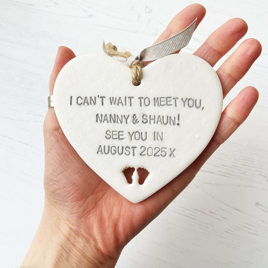 Pearlised white clay hanging heart with baby feet cut out of the bottom and grey personalisation, the heart is personalised with I CAN’T WAIT TO MEET YOU, NANNY & SHAUN! SEE YOU IN AUGUST 2025 X