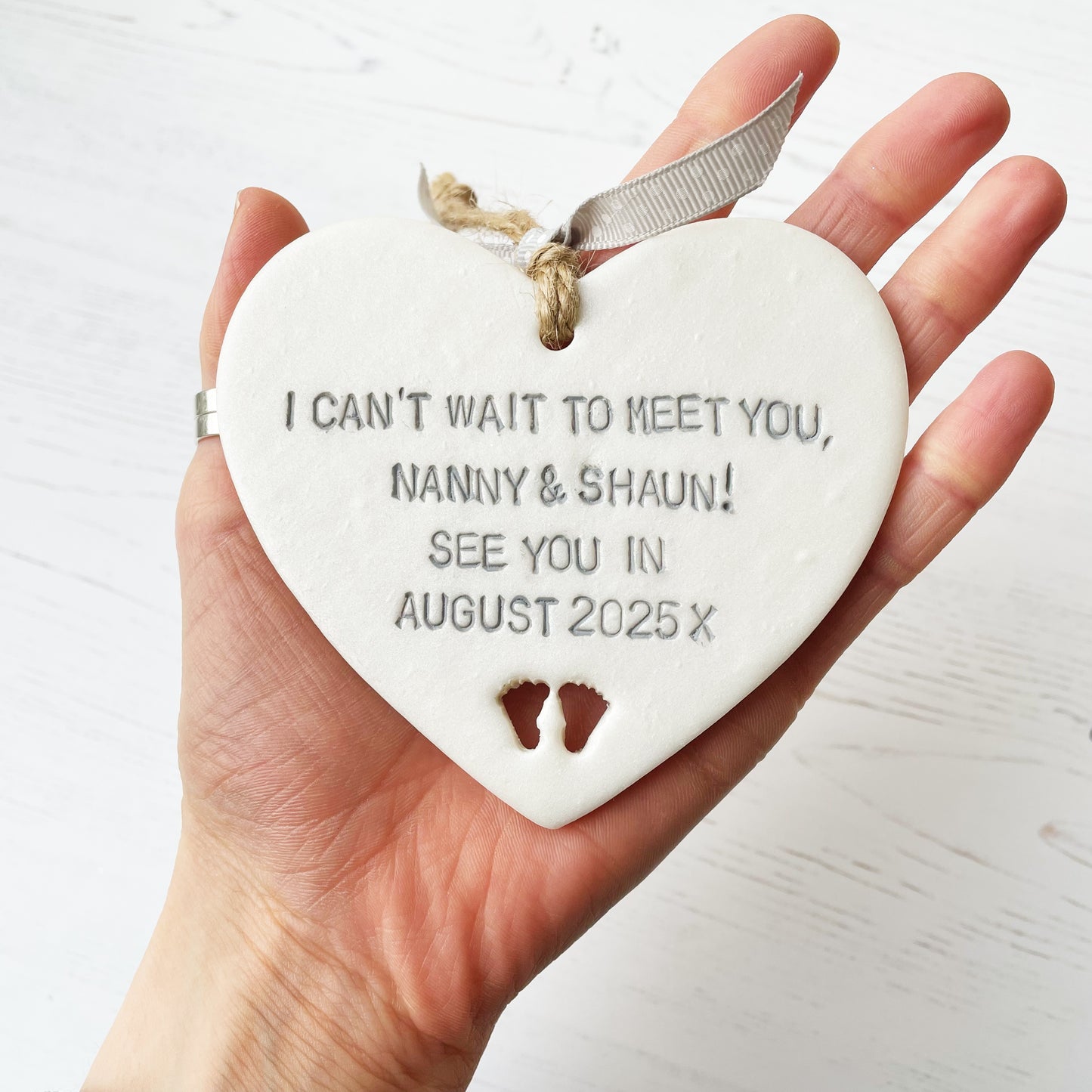 Pearlised white clay hanging heart with baby feet cut out of the bottom and grey personalisation, the heart is personalised with I CAN’T WAIT TO MEET YOU, NANNY! SEE UOU IN AUGUST 2025 X