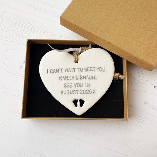 Pearlised white clay hanging heart with baby feet cut out of the bottom and grey personalisation, the heart is personalised with I CAN’T WAIT TO MEET YOU, NANNY & SHAUN! SEE YOU IN AUGUST 2025 X