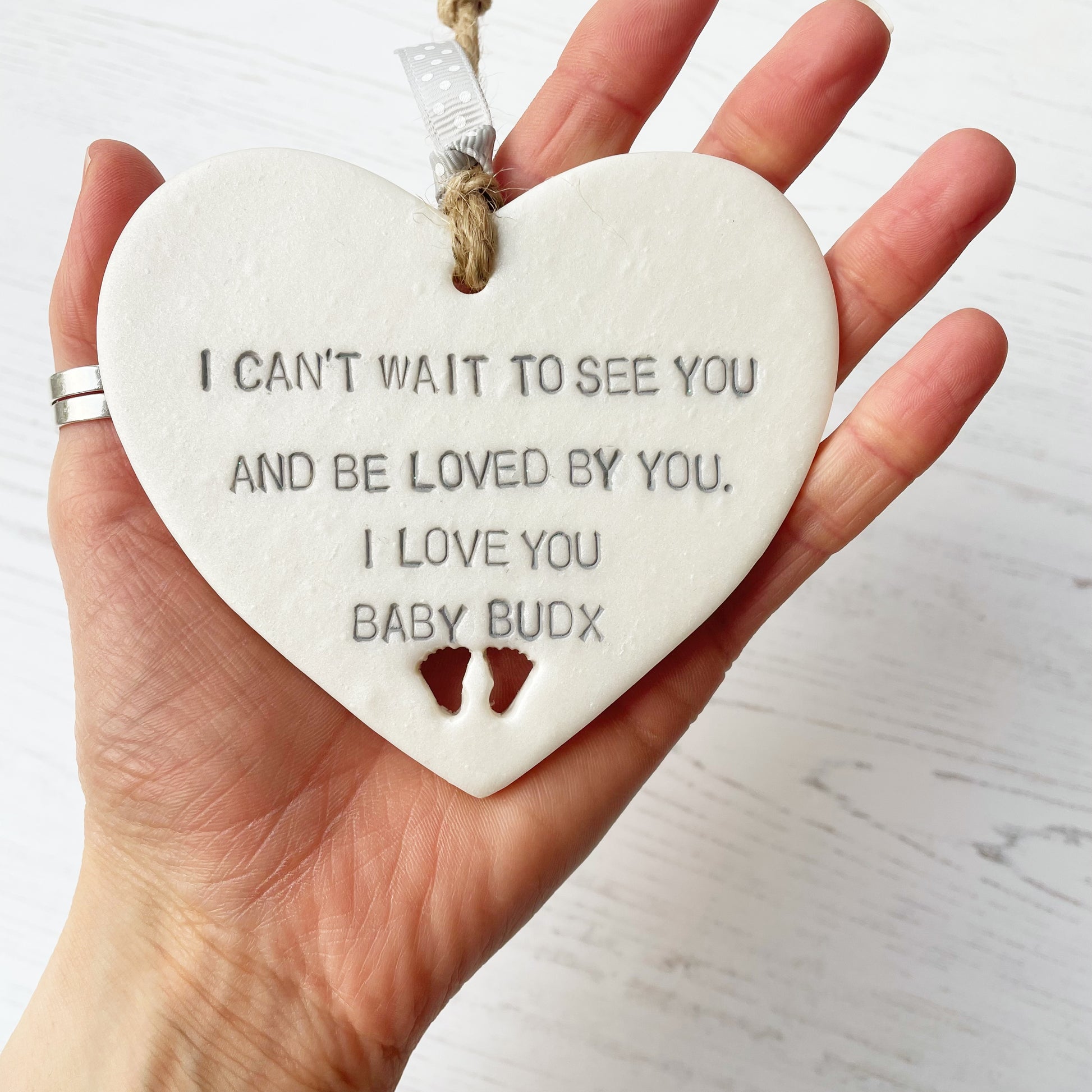 Pearlised white clay hanging heart with baby feet cut out of the bottom and grey personalisation, the heart is personalised with I CAN’T WAIT TO SEE YOU AND BY LOVED BY YOU. I LOVE YOU BABY BUD X