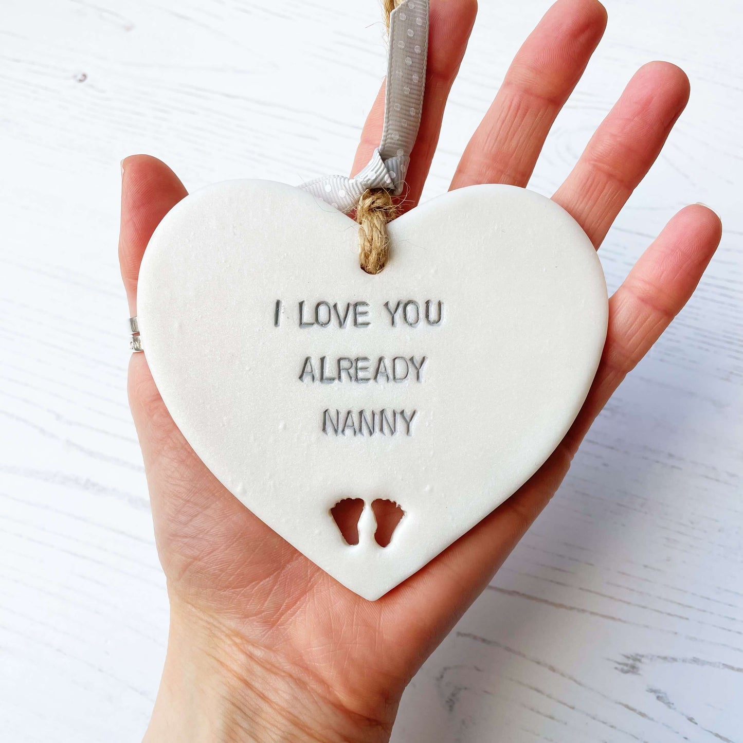 Pearlised white clay hanging heart pregnancy reveal with baby feet cut out of the bottom and grey personalisation, the heart is personalised with I LOVE YOU ALREADY NANNY