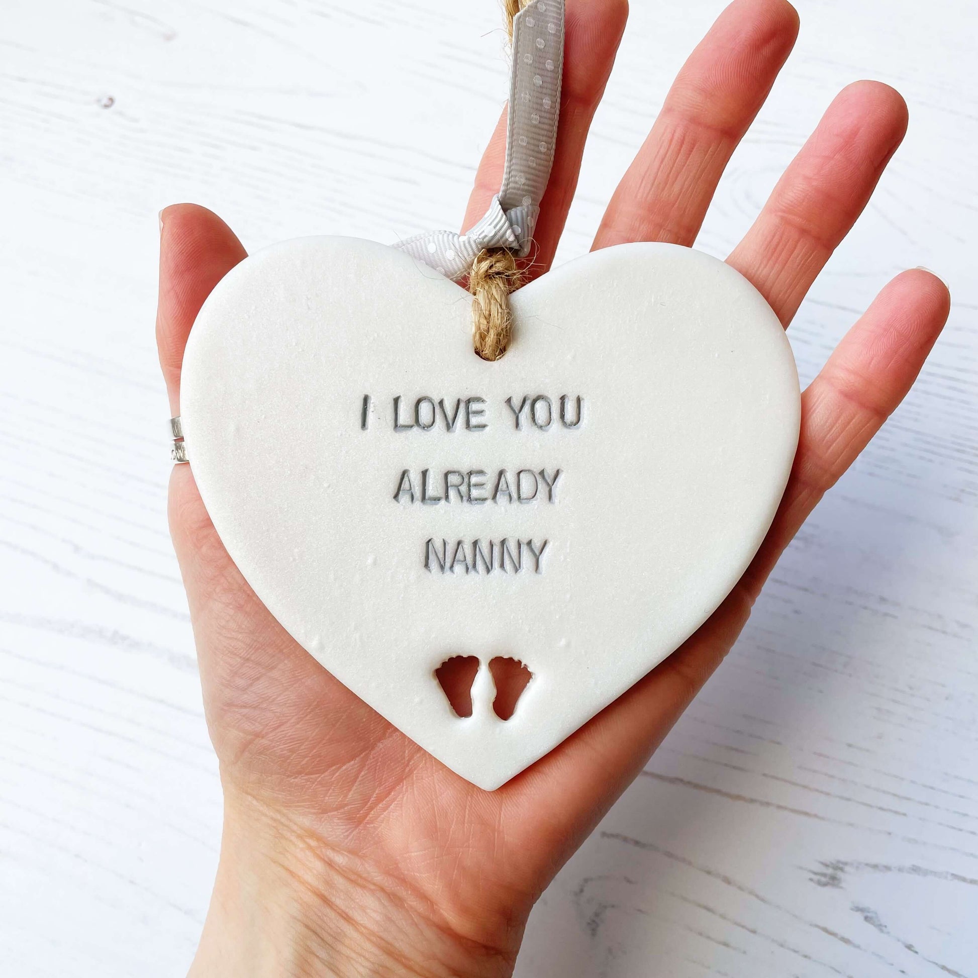 Pearlised white clay hanging heart pregnancy reveal with baby feet cut out of the bottom and grey personalisation, the heart is personalised with I LOVE YOU ALREADY NANNY