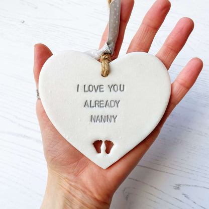 Pearlised white clay hanging heart pregnancy reveal with baby feet cut out of the bottom and grey personalisation, the heart is personalised with I LOVE YOU ALREADY NANNY