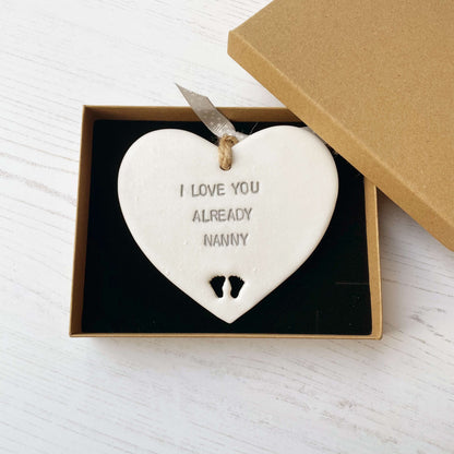 Pearlised white clay hanging heart pregnancy reveal with baby feet cut out of the bottom and grey personalisation, the heart is personalised with I LOVE YOU ALREADY NANNY
