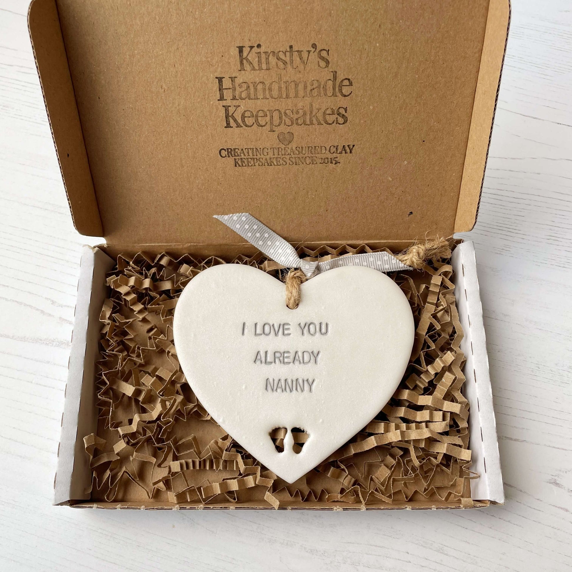 Pearlised white clay hanging heart pregnancy reveal with baby feet cut out of the bottom and grey personalisation, the heart is personalised with I LOVE YOU ALREADY NANNY