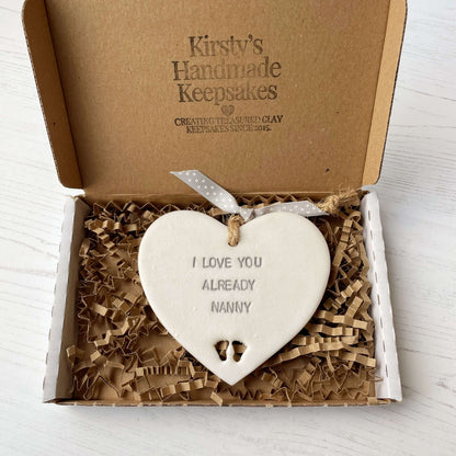 Pearlised white clay hanging heart pregnancy reveal with baby feet cut out of the bottom and grey personalisation, the heart is personalised with I LOVE YOU ALREADY NANNY