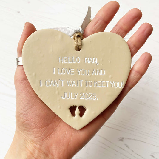 Beige clay hanging heart with baby feet cut out of the bottom and white personalisation, the heart is personalised with HELLO NAN, I LOVE YOU AND I CAN’T WAIT TO MEET YOU! JULY 2025.