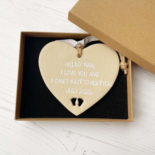 Beige clay hanging heart with baby feet cut out of the bottom and white personalisation, the heart is personalised with HELLO NAN, I LOVE YOU AND I CAN’T WAIT TO MEET YOU! JULY 2025.