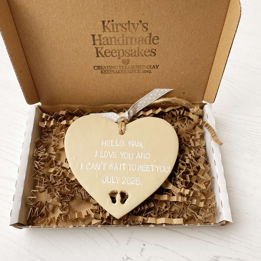Beige clay hanging heart with baby feet cut out of the bottom and white personalisation, the heart is personalised with HELLO NAN, I LOVE YOU AND I CAN’T WAIT TO MEET YOU! JULY 2025.