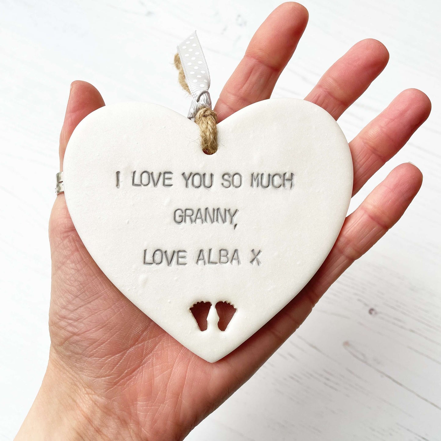 Pearlised white clay hanging heart with baby feet cut out of the bottom and grey personalisation, the heart is personalised with I LOVE YOU SO MUCH GRANNY, LOVE ALBA X