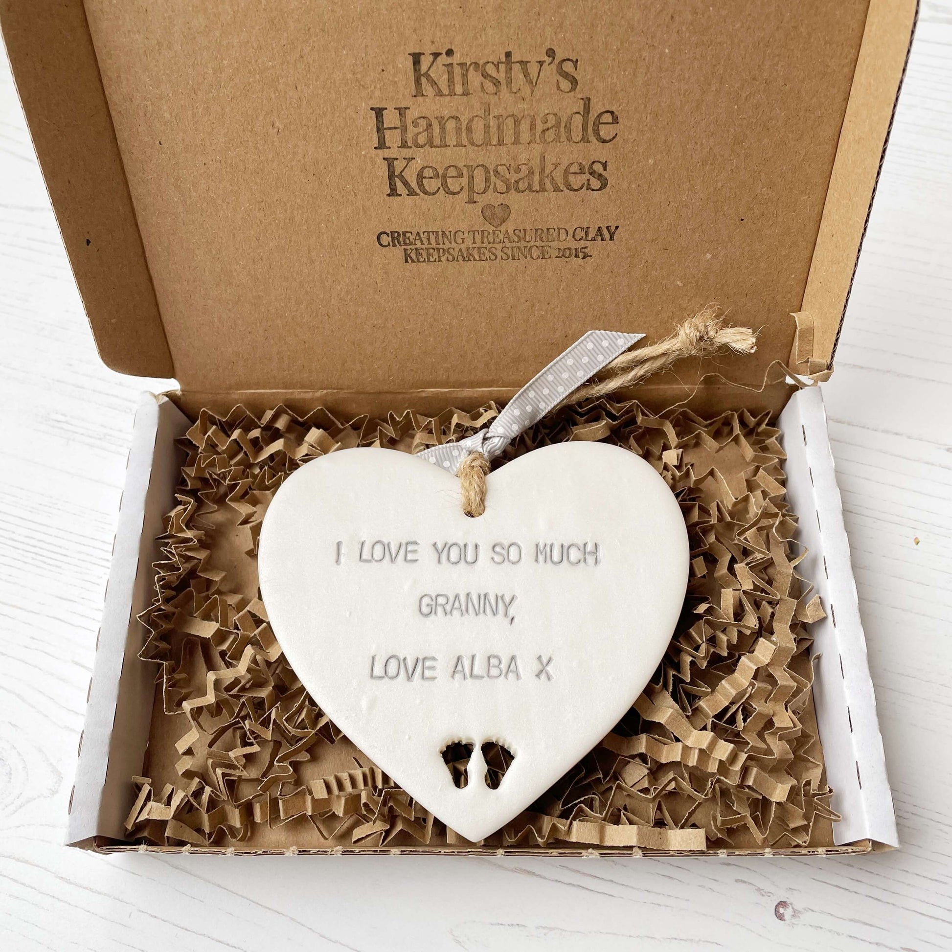 Pearlised white clay hanging heart with baby feet cut out of the bottom and grey personalisation, the heart is personalised with I LOVE YOU SO MUCH GRANNY, LOVE ALBA X