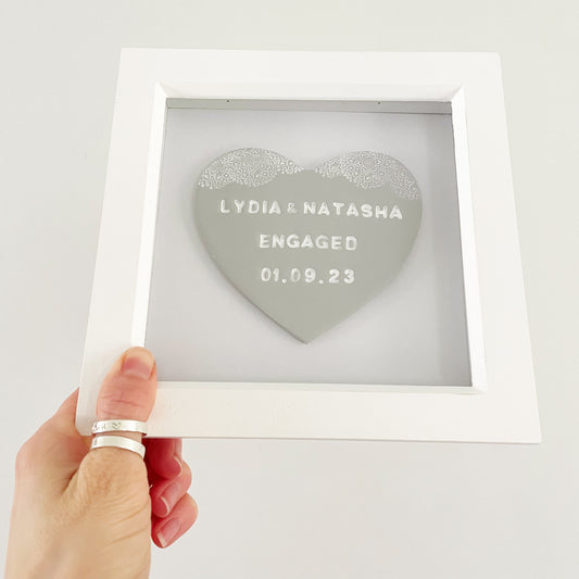 Personalised framed lesbian engagement gift, grey clay heart with a white lace edge at the top of the heart in a white box frame, the heart is personalised with LYDIA & NATASHA ENGAGED 01.09.23