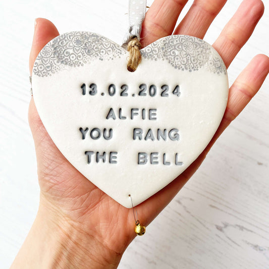 Personalised cancer survivor gift, pearlised white clay hanging heart with a grey lace edge at the top of the heart and a gold bell hanging below, the heart is personalised with 13.02.2024 ALFIE YOU RANG THE BELL