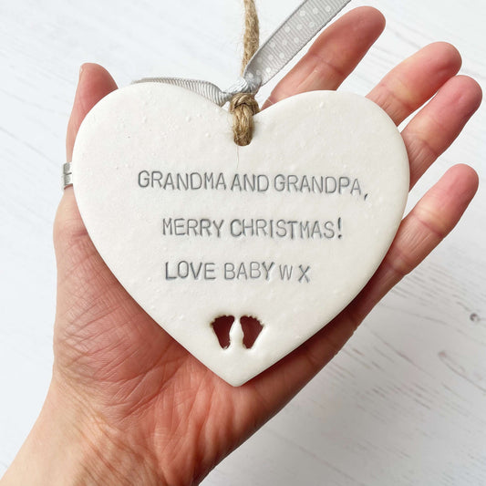 Pearlised white clay hanging heart with baby feet cut out of the bottom and grey personalisation, the heart is personalised with GRANDMA & GRANDPA, MERRY CHRISTMAS! LOVE BABY W X