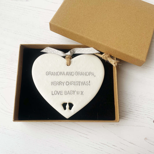 Pearlised white clay hanging heart with baby feet cut out of the bottom and grey personalisation, the heart is personalised with GRANDMA & GRANDPA, MERRY CHRISTMAS! LOVE BABY W X