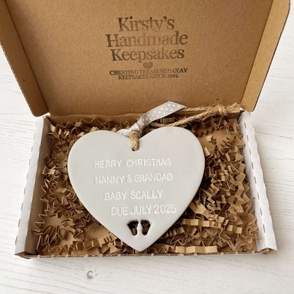Grey clay hanging heart with baby feet cut out of the bottom and white personalisation, the heart is personalised with MERRY CHRISTMAS NANNY & GRANDAD BABY SCALLY DUE JULY 2025