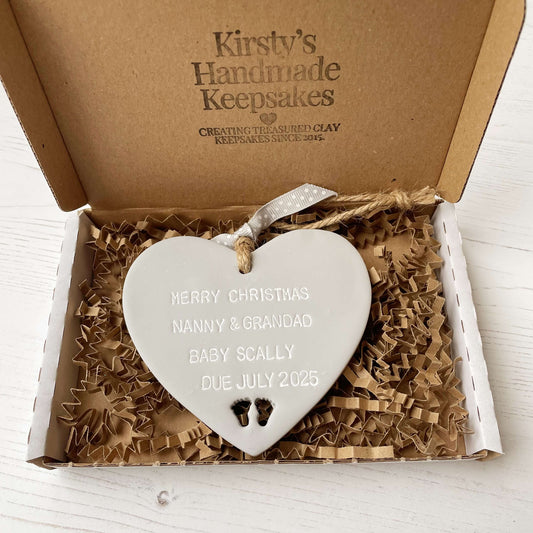 Grey clay hanging heart with baby feet cut out of the bottom and white personalisation, the heart is personalised with MERRY CHRISTMAS NANNY & GRANDAD BABY SCALLY DUE JULY 2025