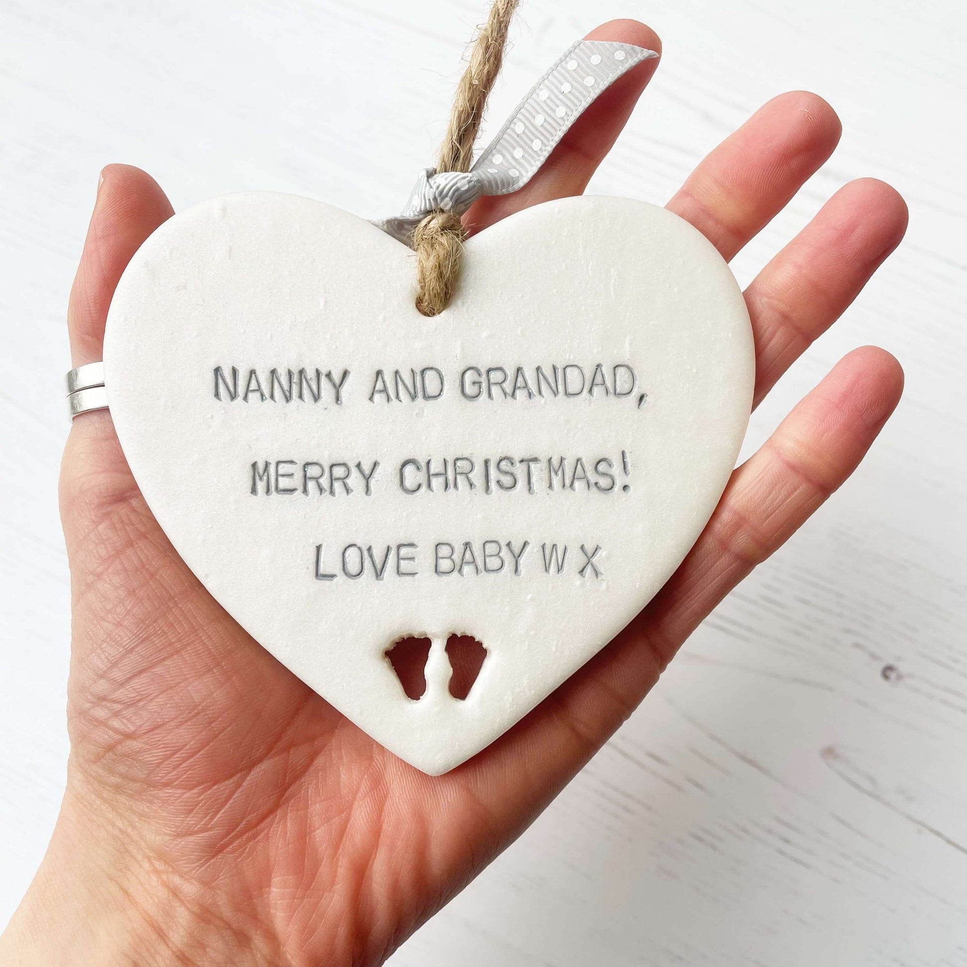 Pearlised white clay hanging heart with baby feet cut out of the bottom and grey personalisation, the heart is personalised with NANNY & GRANDAD, MERRY CHRISTMAS! LOVE BABY W X