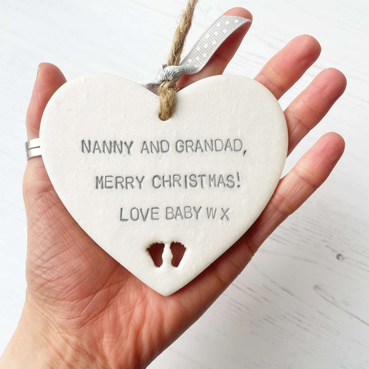 Pearlised white clay hanging heart with baby feet cut out of the bottom and grey personalisation, the heart is personalised with NANNY & GRANDAD, MERRY CHRISTMAS! LOVE BABY W X