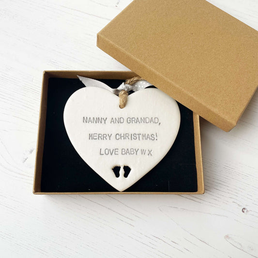 Pearlised white clay hanging heart with baby feet cut out of the bottom and grey personalisation, the heart is personalised with NANNY & GRANDAD, MERRY CHRISTMAS! LOVE BABY W X