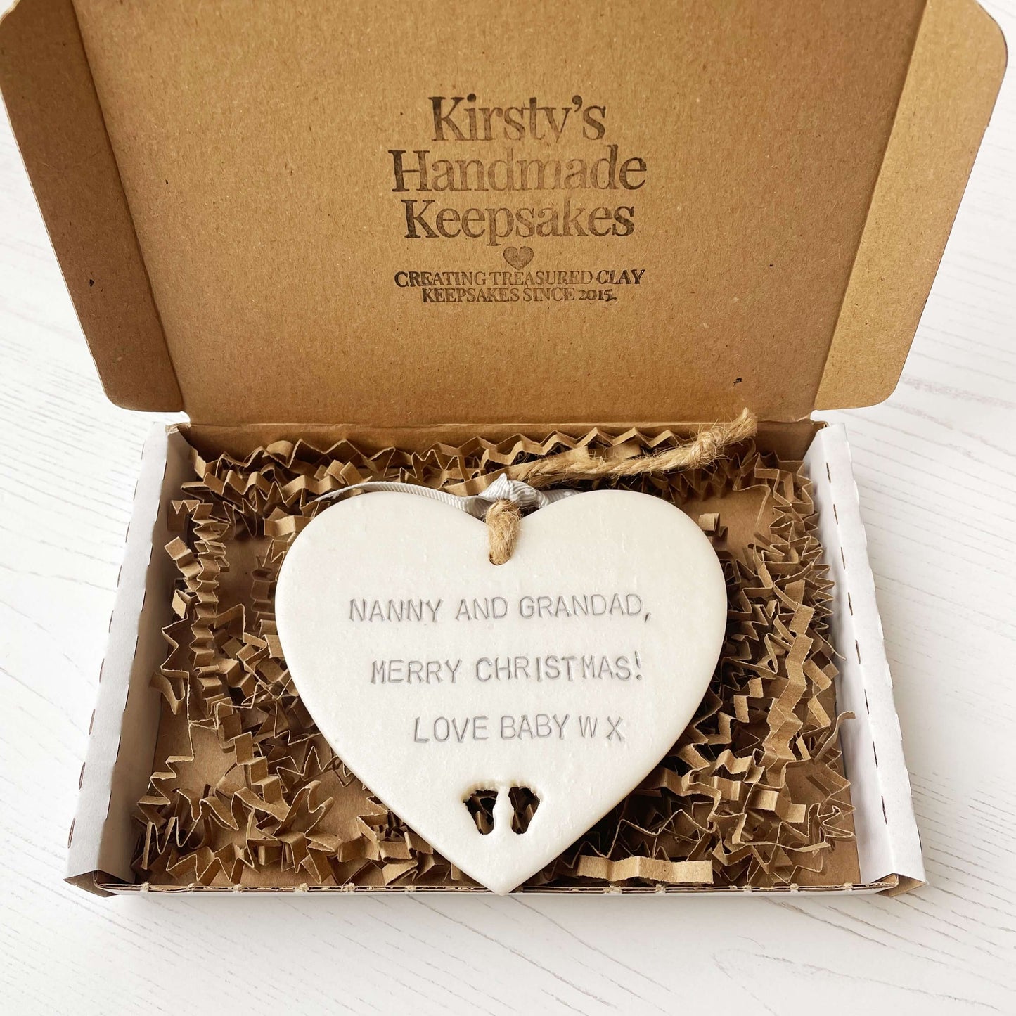 Pearlised white clay hanging heart with baby feet cut out of the bottom and grey personalisation, the heart is personalised with NANNY & GRANDAD, MERRY CHRISTMAS! LOVE BABY W X