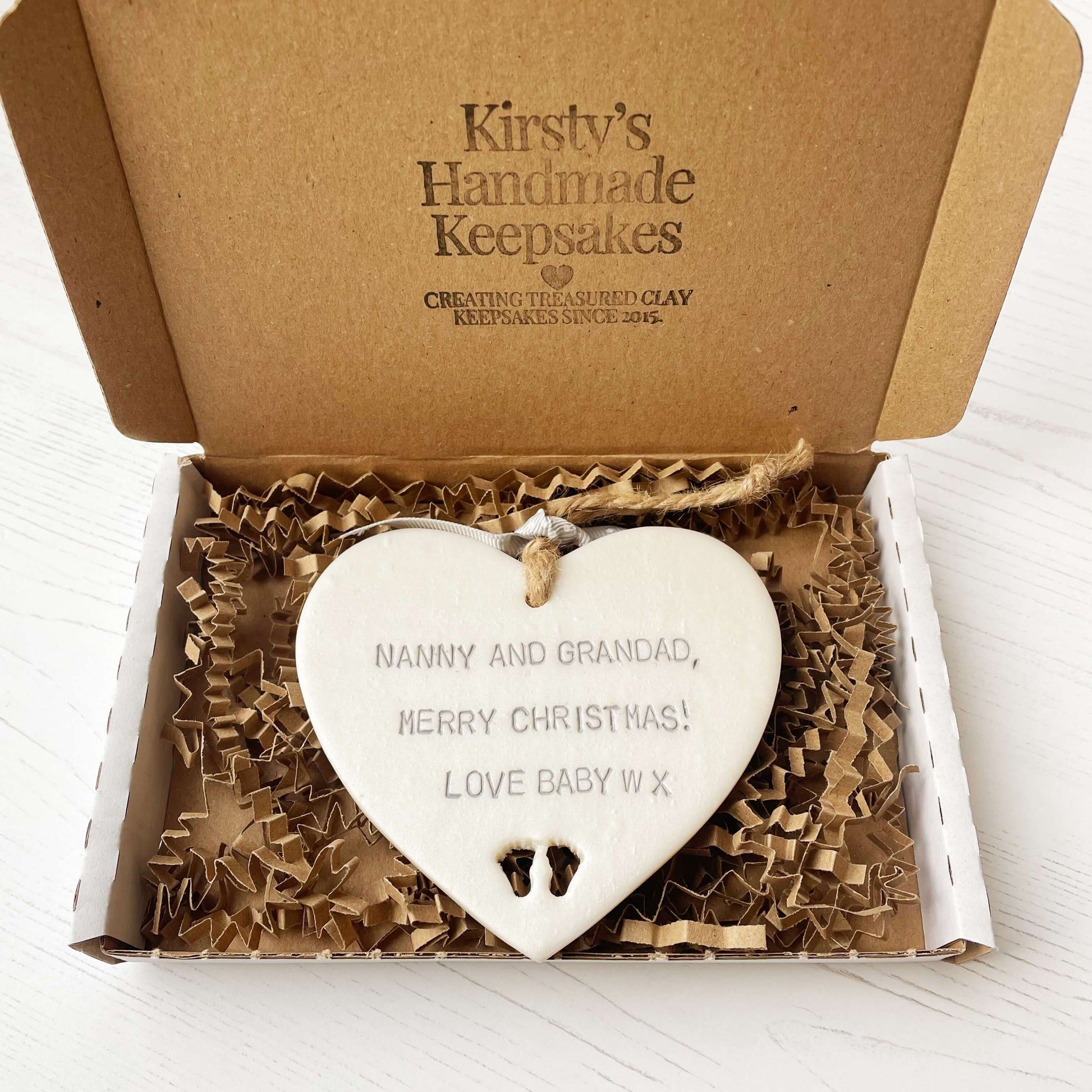 Pearlised white clay hanging heart with baby feet cut out of the bottom and grey personalisation, the heart is personalised with NANNY & GRANDAD, MERRY CHRISTMAS! LOVE BABY W X