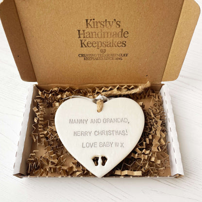 Pearlised white clay hanging heart with baby feet cut out of the bottom and grey personalisation, the heart is personalised with NANNY & GRANDAD, MERRY CHRISTMAS! LOVE BABY W X