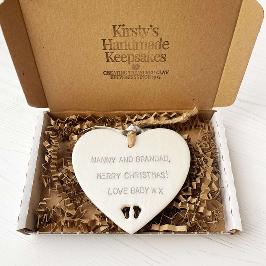 Pearlised white clay hanging heart with baby feet cut out of the bottom and grey personalisation, the heart is personalised with NANNY & GRANDAD, MERRY CHRISTMAS! LOVE BABY W X