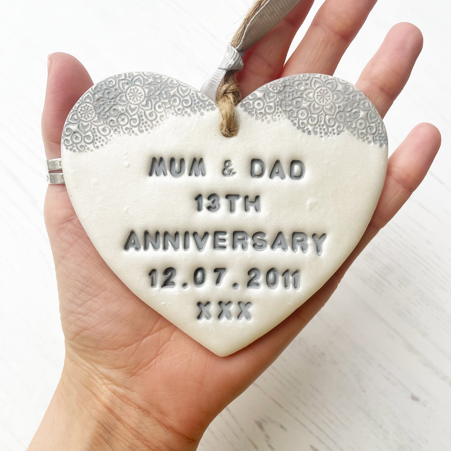 Personalised 13th anniversary gift, pearlised white clay hanging heart with a grey lace edge at the top of the heart, the heart is personalised with MUM & DAD 13TH ANNIVERSARY 12.07.2011 XXX