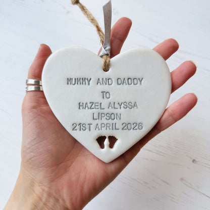 Pearlised white clay hanging heart with baby feet cut out of the bottom and grey personalisation, the heart is personalised with MUMMY & DADDY TO HAZEL ALYSSA LIPSON 21ST APRIL 2026